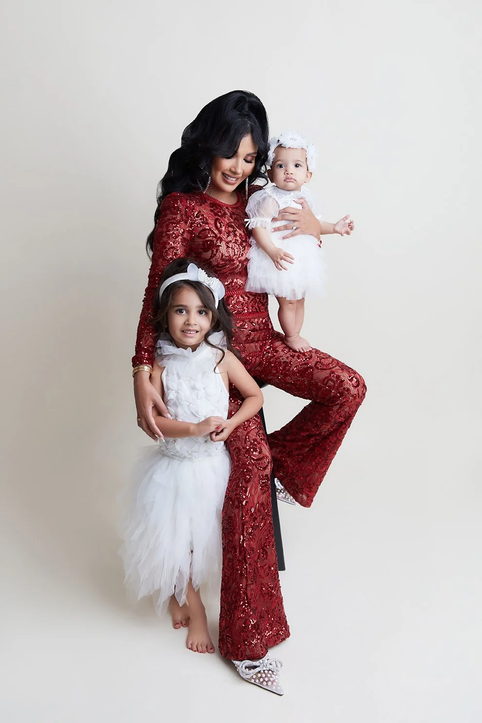 A woman in a red sequined outfit sitting on a stool with two young girls in white dresses, against a plain light background.