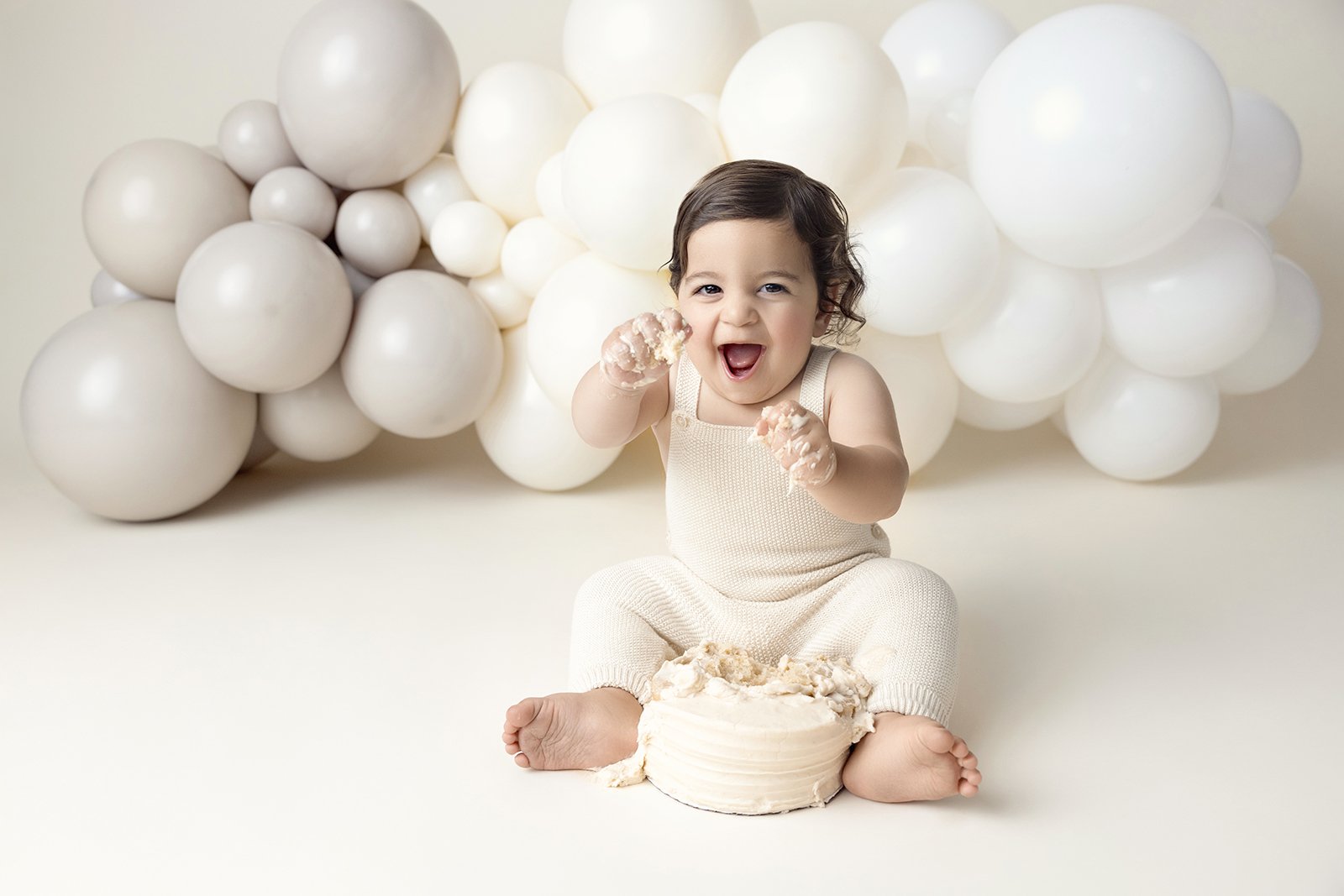 A young child sitting on the floor, smiling and playing with a cake, with white balloons in the background.
