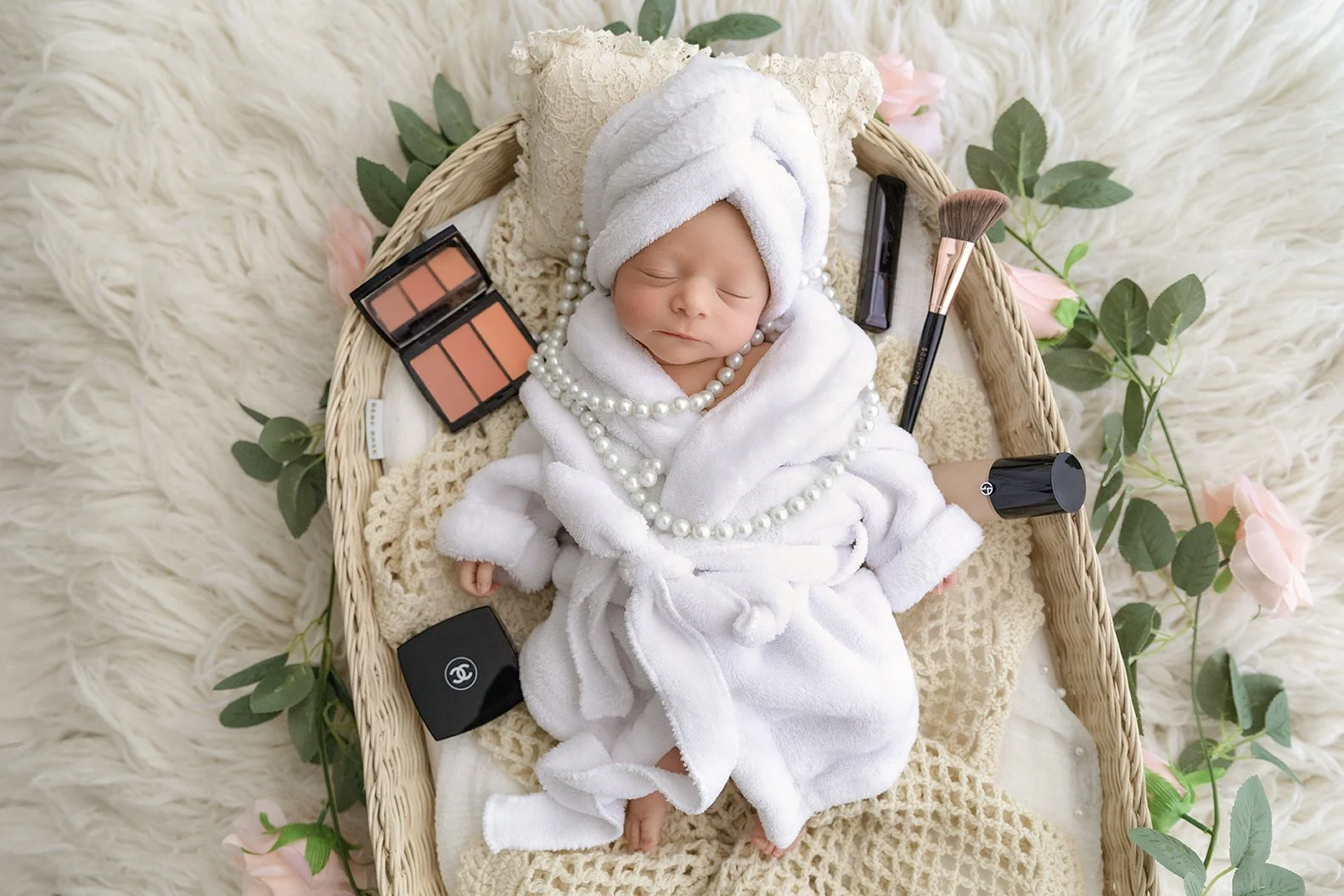 A baby is sleeping in a woven basket, dressed in a white bathrobe and towel wrapped around their head. The basket is lined with beige and cream blankets, surrounded by pink roses and green leaves. Various makeup items, including blush, lipstick, and 