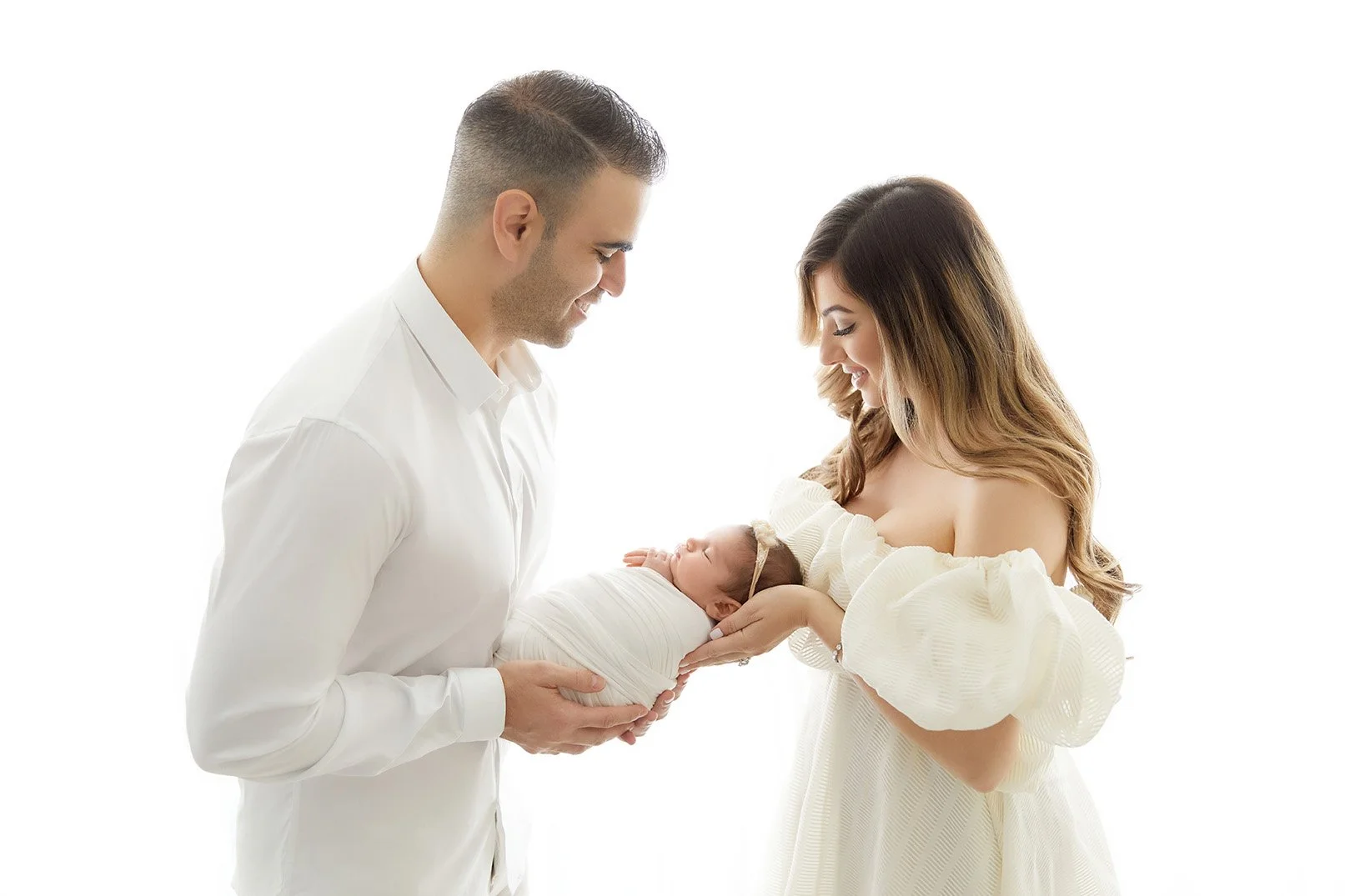 A couple holding a newborn baby, looking at each other and smiling, against a white background.