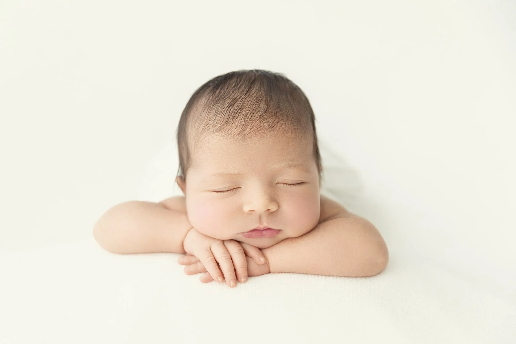 Close-up of a sleeping newborn baby with hands tucked under chin, lying on a white surface.