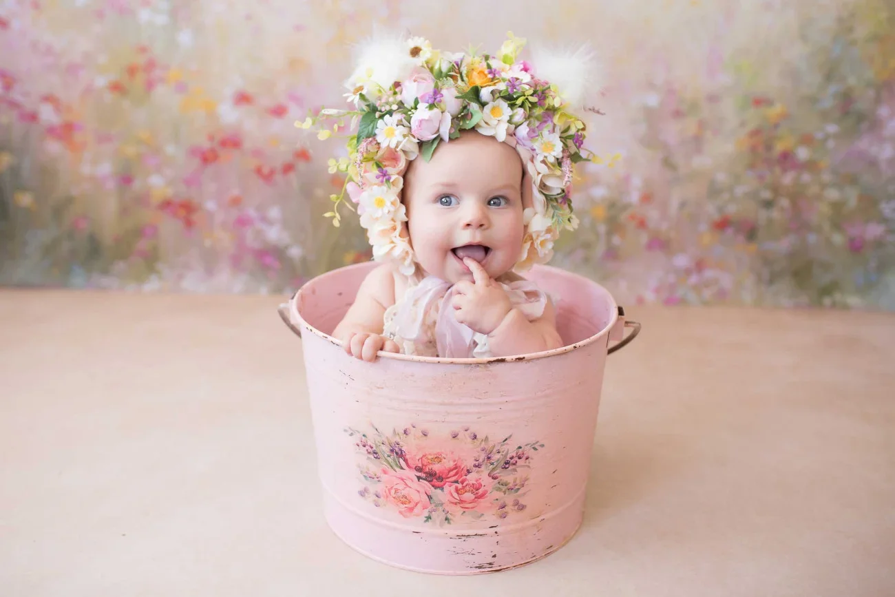 Baby sitting in a pink vintage bucket, wearing a floral crown with pink, white, and purple flowers, surrounded by a colorful floral background.