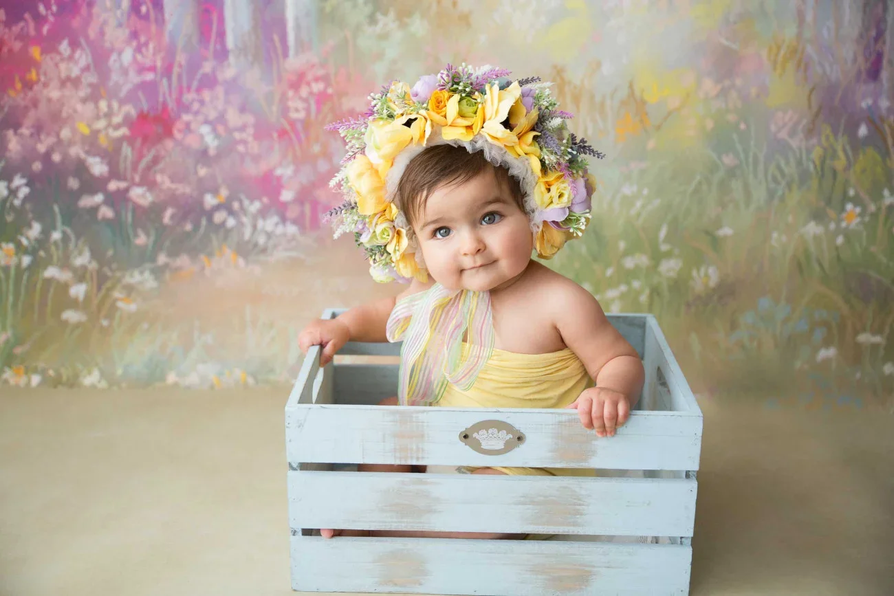 A baby girl wearing a flower crown and a yellow dress, sitting in a white wooden crate, with a colorful floral background.