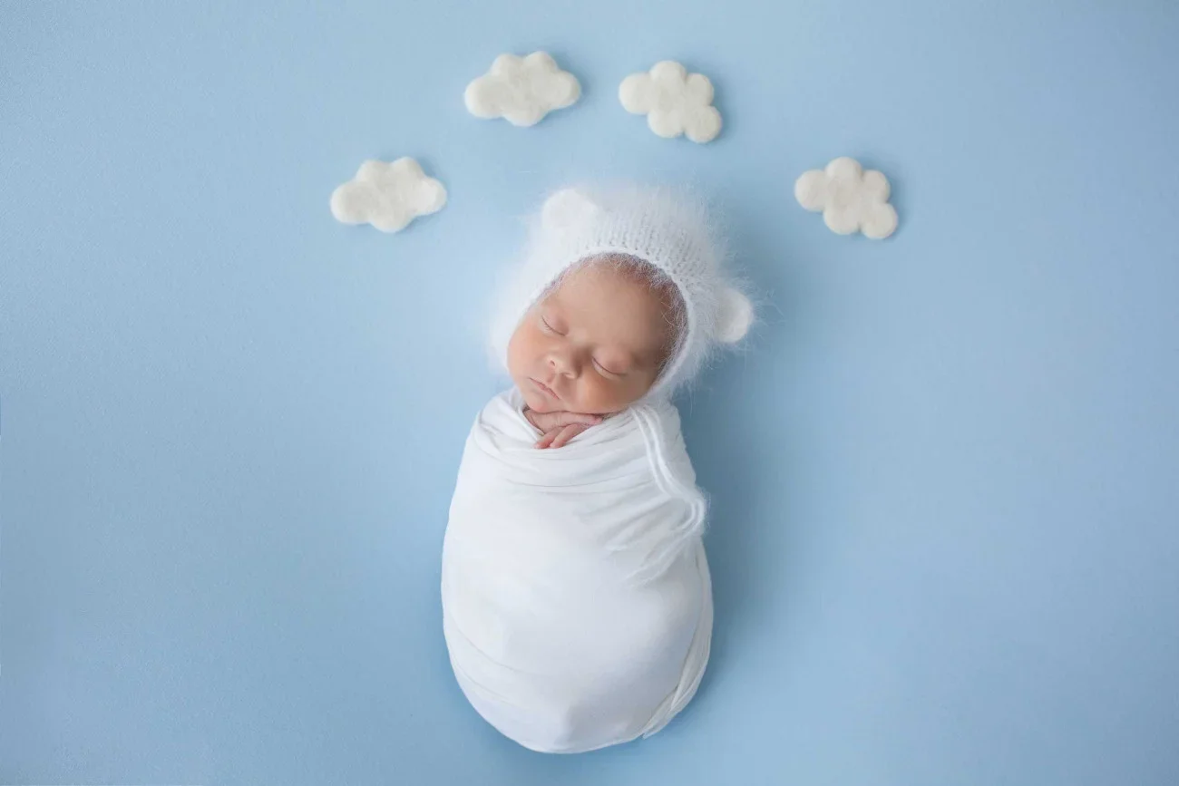 Baby swaddled in white blanket, wearing a fuzzy white hat, sleeping on a light blue background with small white cloud-shaped decorations.