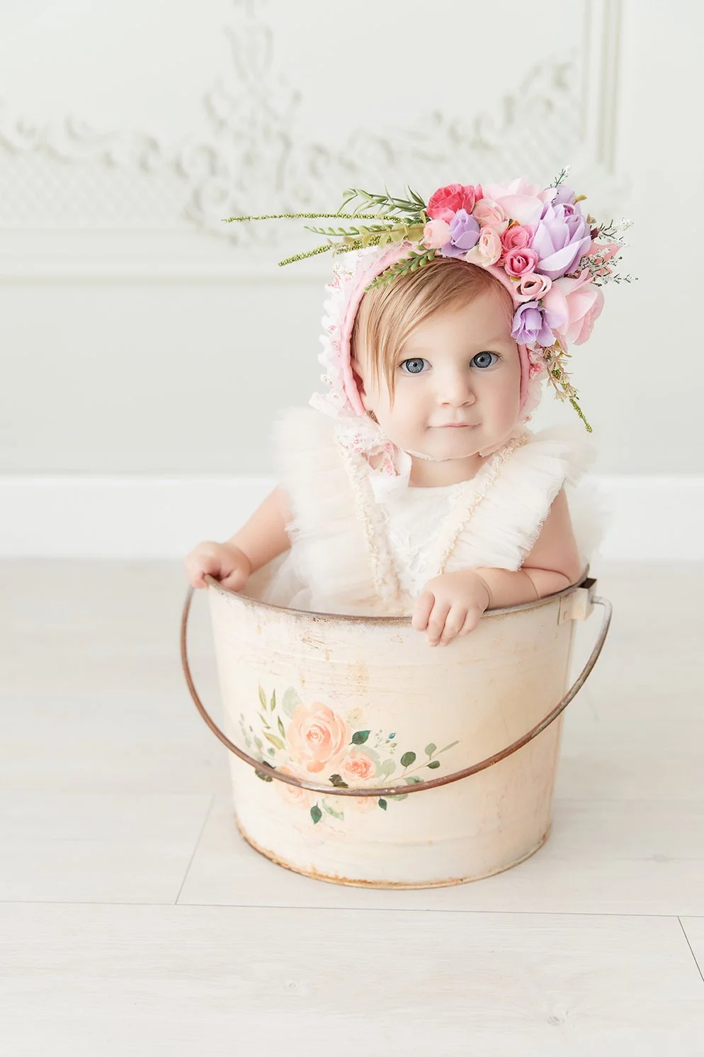 Cute baby girl with gray eyes, wearing a pink floral crown, leaning on a vintage white flower-printed bucket, in a bright room with white walls.