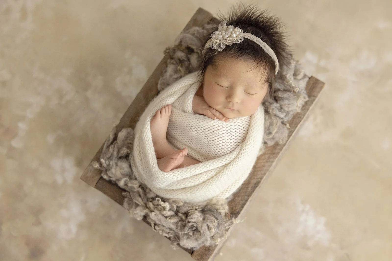 A sleeping newborn baby wrapped in a cream knitted blanket, lying on a small wooden bed with a fluffy gray blanket, wearing a headband with a decorative flower, on a beige surface.
