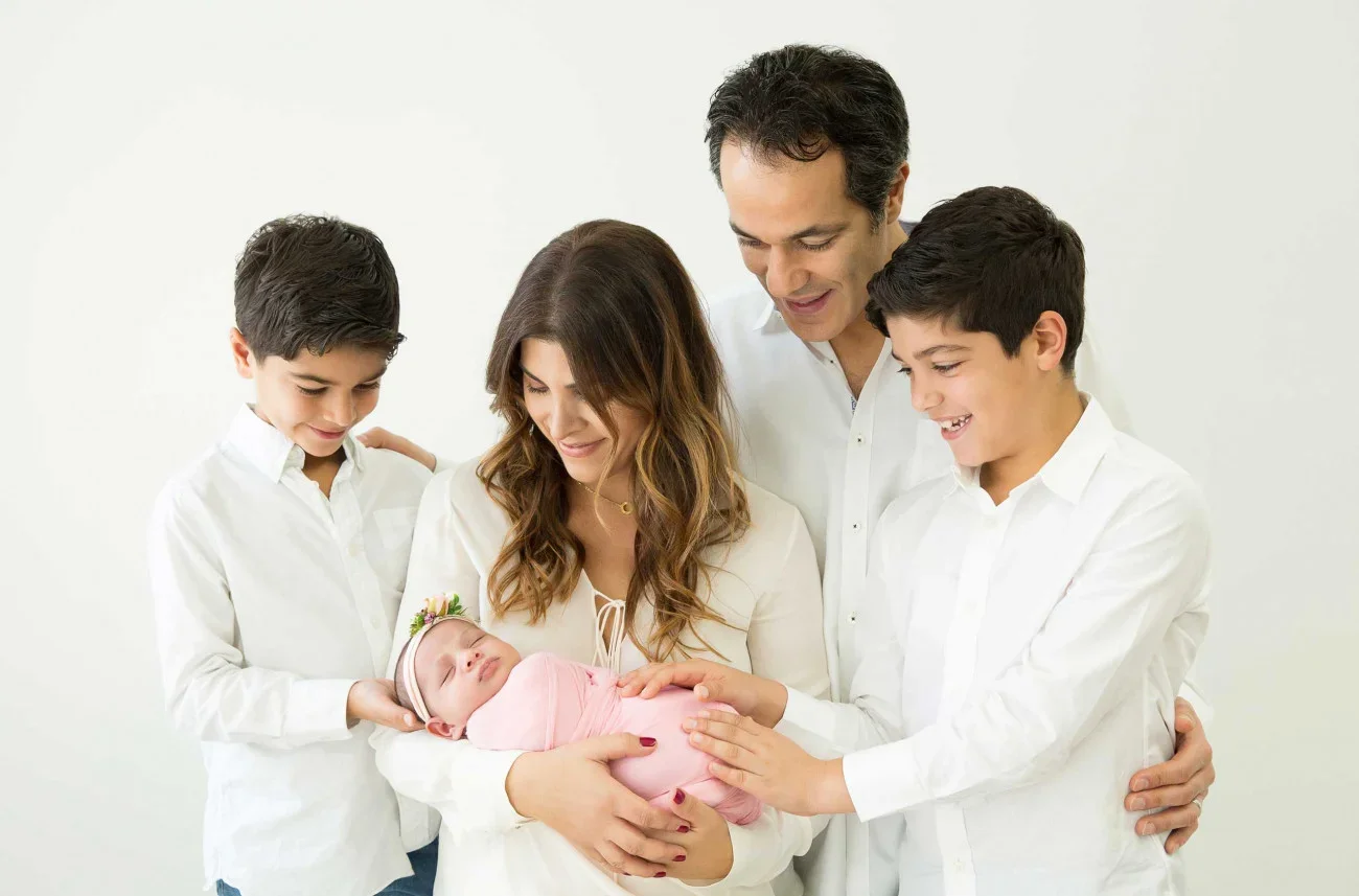 Family with parents and two young boys holding a newborn baby girl wrapped in pink, celebrating together.