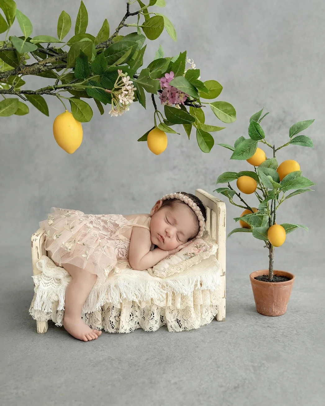 A sleeping baby girl in a lace dress, lying on a small vintage bed, with a headband, surrounded by artificial lemon and orange trees with blossoms, against a plain gray background.