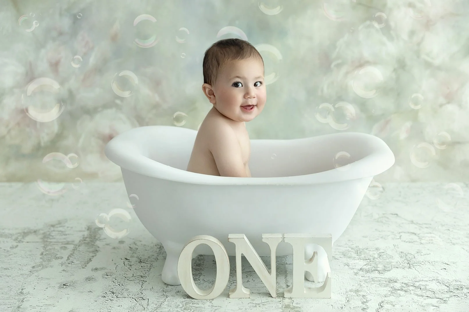 A smiling baby sitting in a white bathtub with a decorative "ONE" sign in front, celebrating their first birthday, surrounded by a soft, pastel-colored background with floating bubbles.