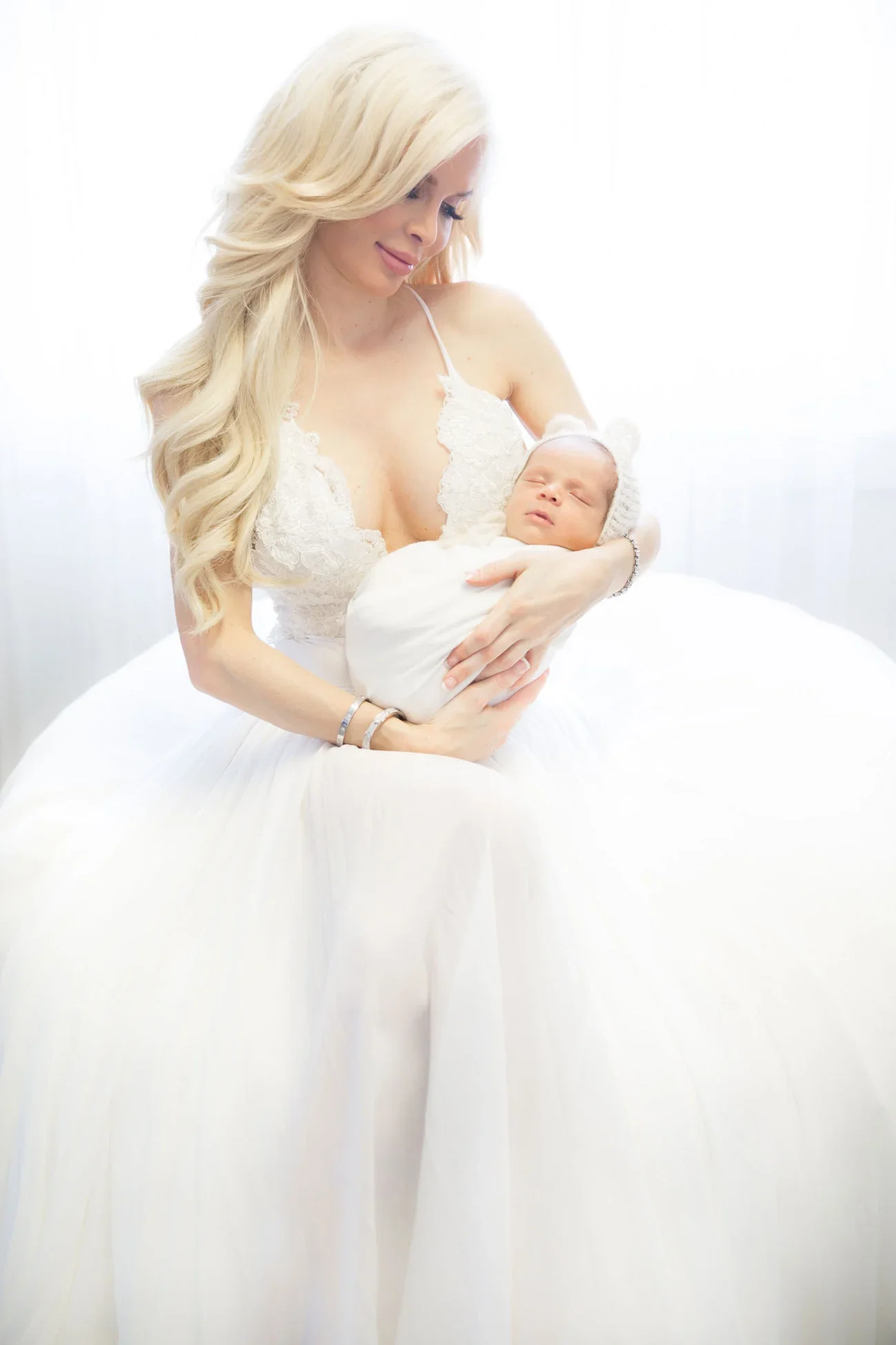 A blonde woman in a white lace dress holds a swaddled baby in her arms, sitting on a white surface near a bright window.