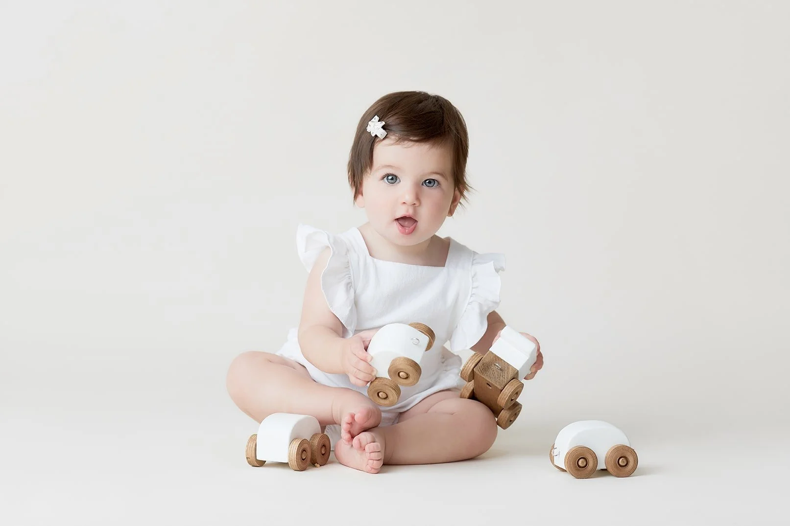 A baby girl with brown hair and blue eyes, wearing a white dress, sitting on a plain white background, playing with wooden and white toy cars.