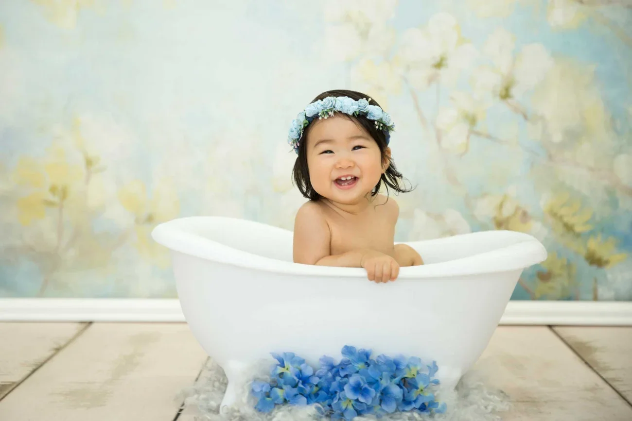 A smiling baby girl sitting in a white bathtub with blue flowers on the floor and wearing a floral headband.