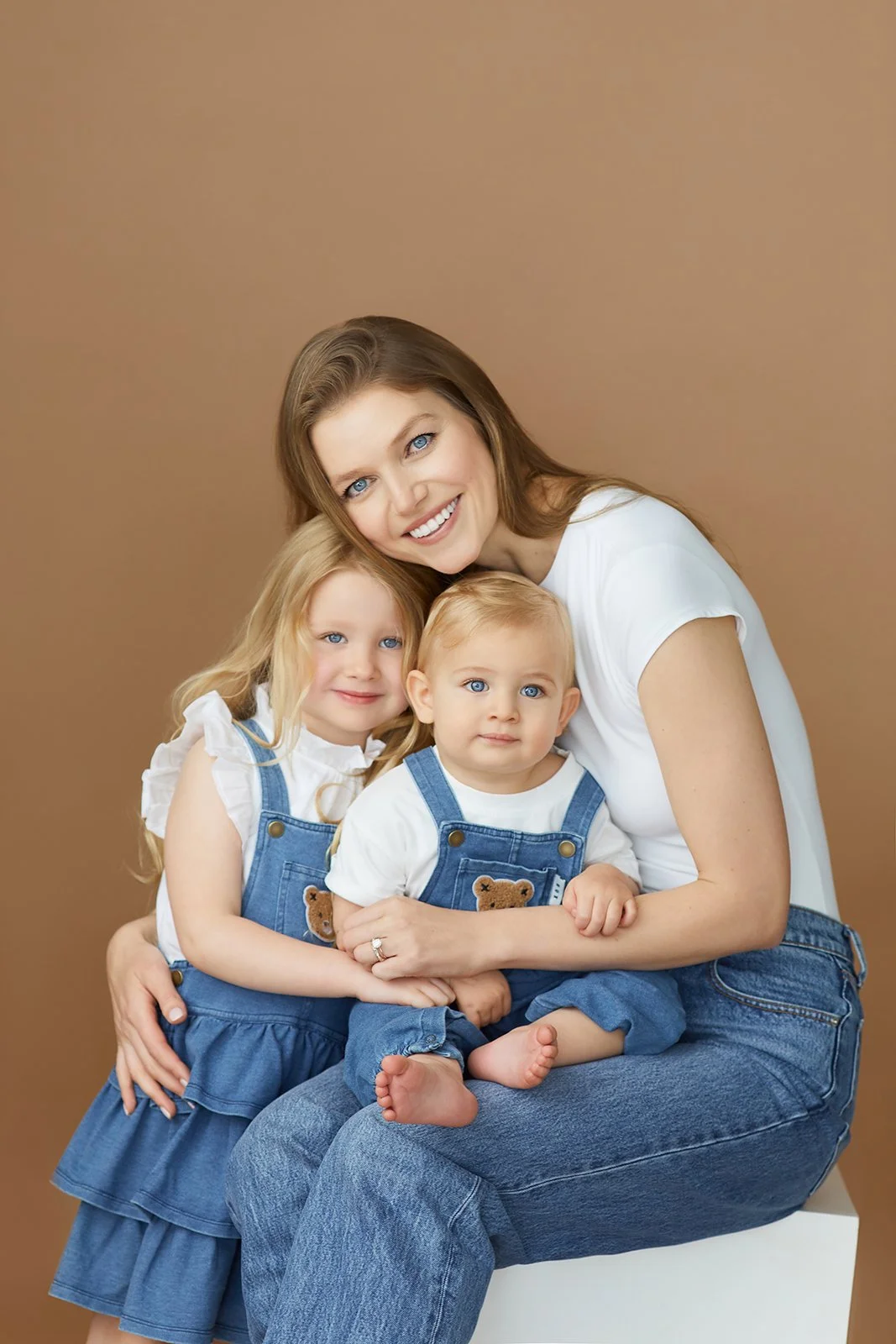 A woman with long brown hair, wearing a white shirt, smiling and hugging two young children with blonde hair and blue eyes, wearing coordinating white shirts and denim outfits, seated against a plain brown background.