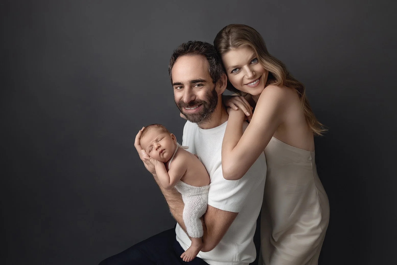 A happy family of three, including a man, a woman, and a sleeping baby, posing together against a dark gray background.