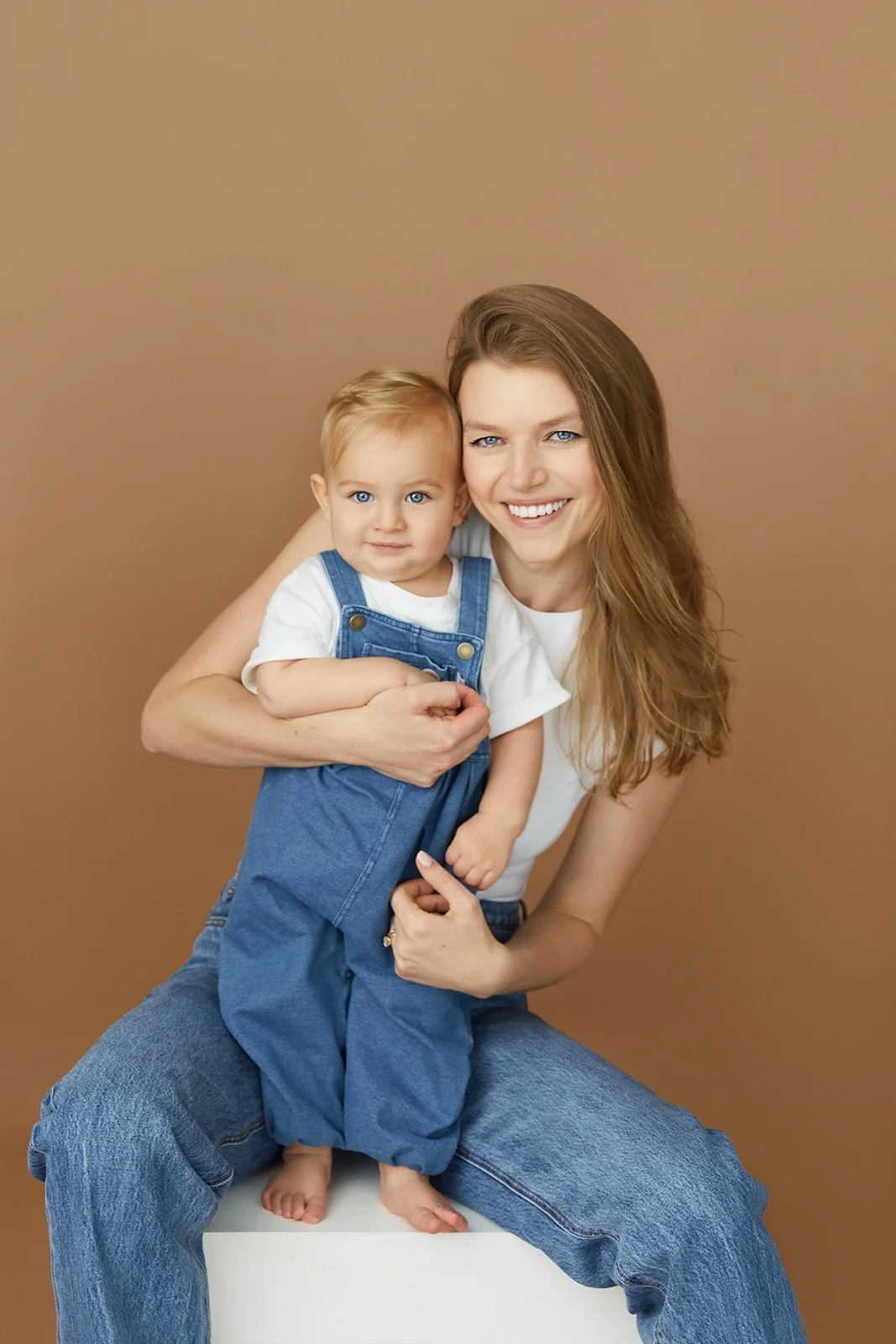 A smiling young woman with long wavy hair holding a toddler boy with blonde hair and blue eyes, against a plain brown background.