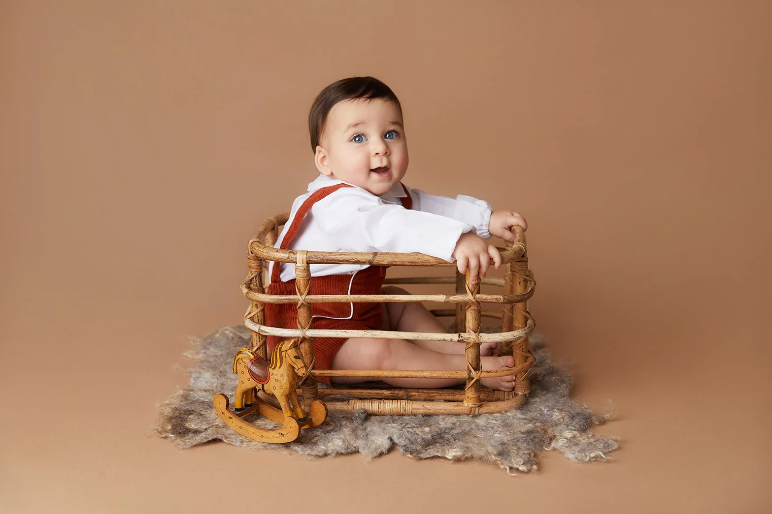 A cute baby with brown hair and blue eyes sitting in a wicker basket, holding the edge, with a wooden rocking horse toy nearby, on a textured rug against a plain brown background.