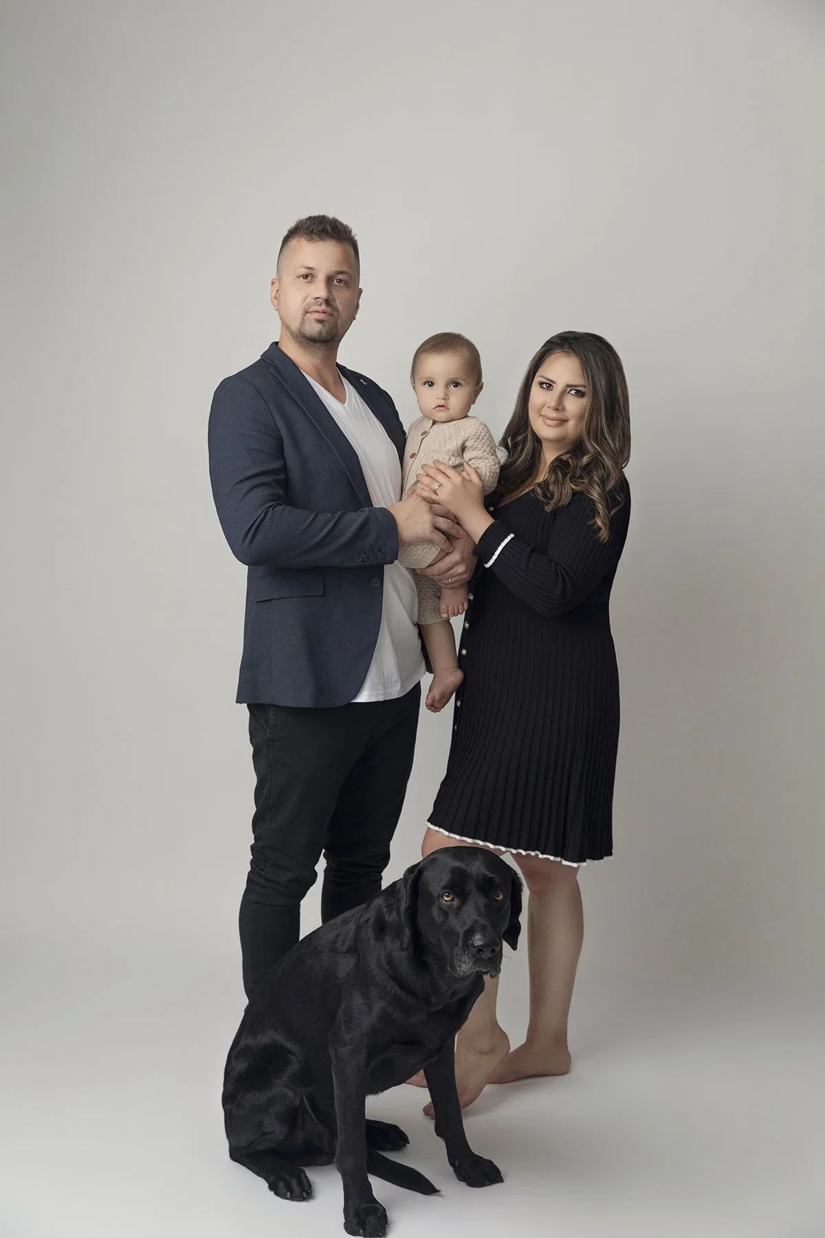 Family portrait with a man, woman, a baby, a dog, and a plain light background.
