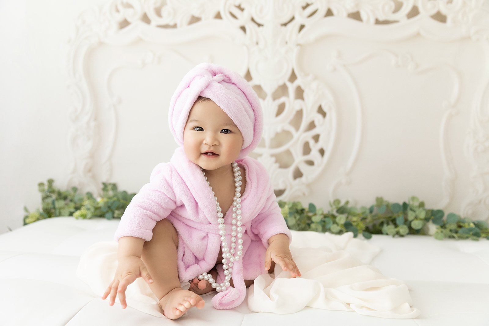 A happy baby girl wearing a pink towel wrapping her hair, pink robe, and a pearl necklace sitting on a bed with white sheets, in a bright decorated room with a white ornate headboard and greenery in the background.