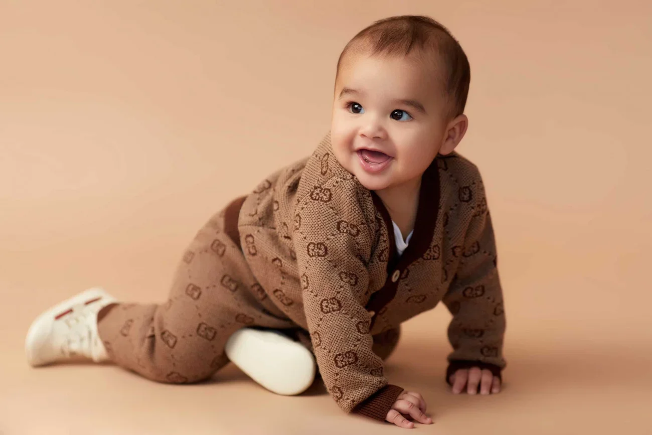 Baby crawling on a tan background, wearing a matching brown Gucci pajamas and white sneakers.