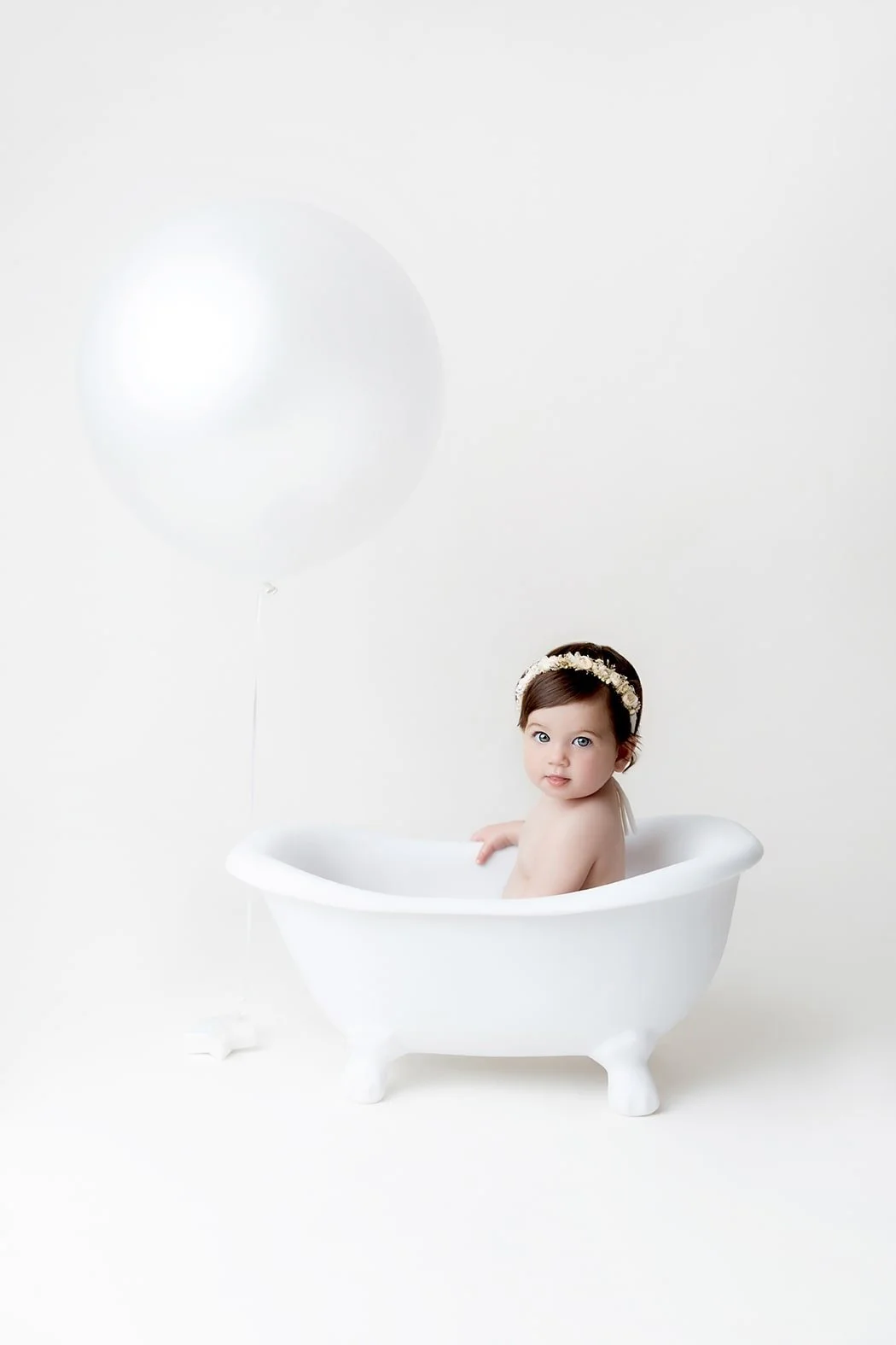 A baby sitting in a small white bathtub against a white background, with a white balloon floating nearby and wearing a floral headband.