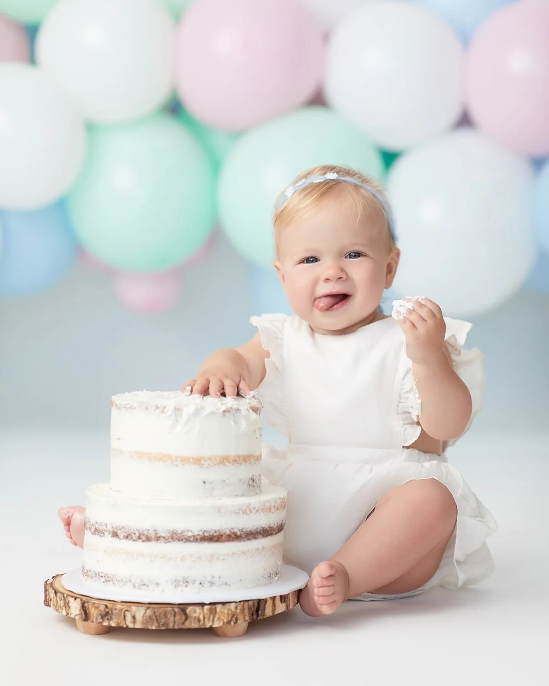 A smiling baby girl with light hair and a white headband, sitting next to a two-tiered birthday cake with white frosting on a wooden cake stand, in front of pastel-colored balloons, celebrating a birthday.