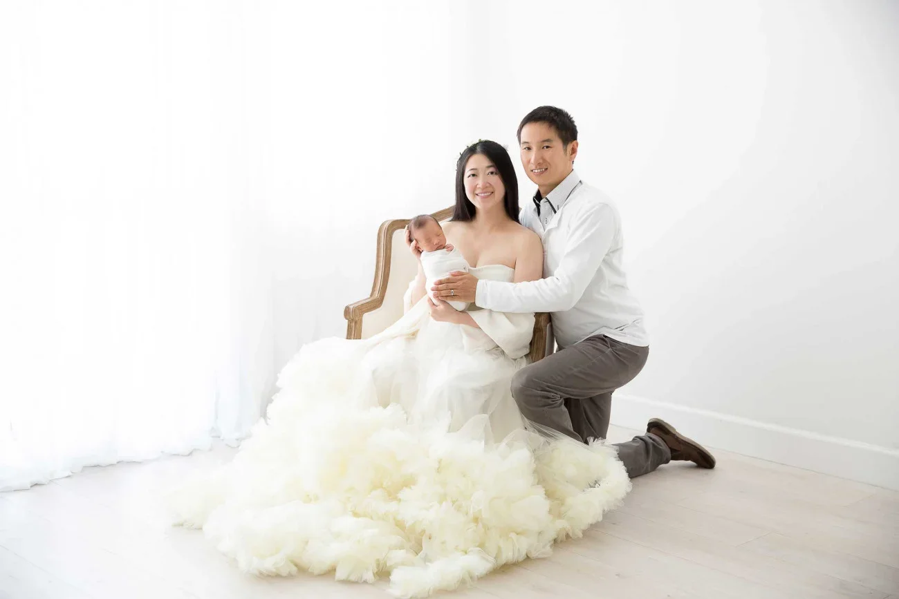 A family photo with a woman in a white dress holding a baby, a man kneeling beside her, all smiling in a bright, minimalist room.