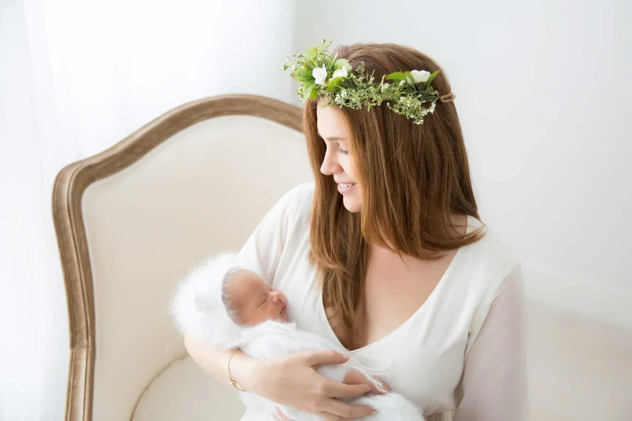 A woman with long brown hair wearing a flower crown, holding a newborn baby wrapped in a white blanket in a bright room.