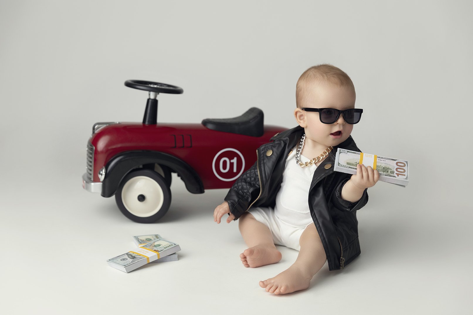 Baby sitting on the floor wearing sunglasses and a leather jacket, holding a stack of cash with a toy car in the background.