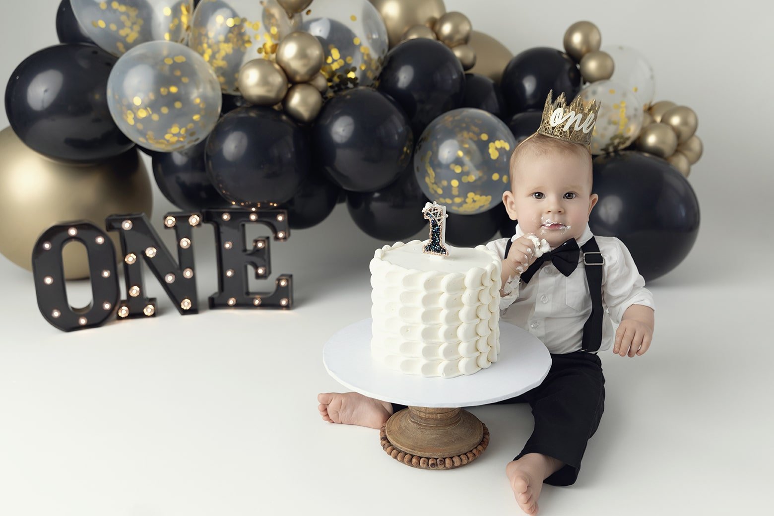 Baby celebrating first birthday with a cake, wearing a gold crown that says 'one,' surrounded by black, gold, and transparent balloons, and illuminated 'ONE' sign in the background.