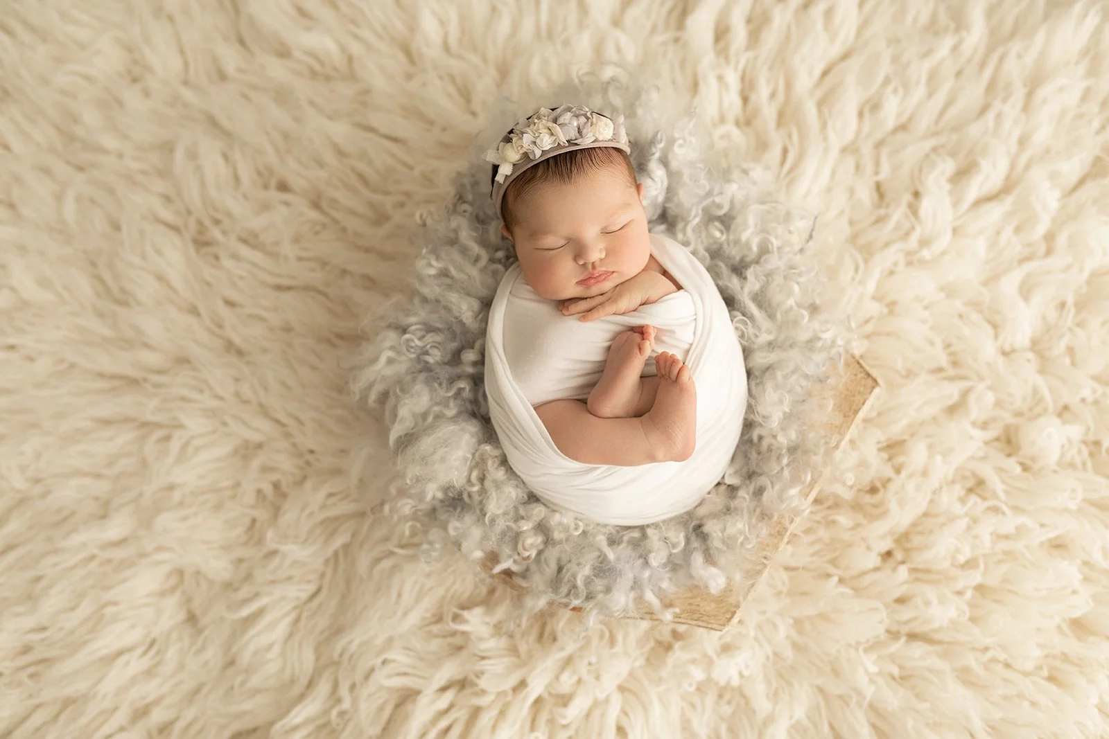 A newborn baby sleeping peacefully swaddled in white, with a floral headband, on a fluffy cream-colored blanket