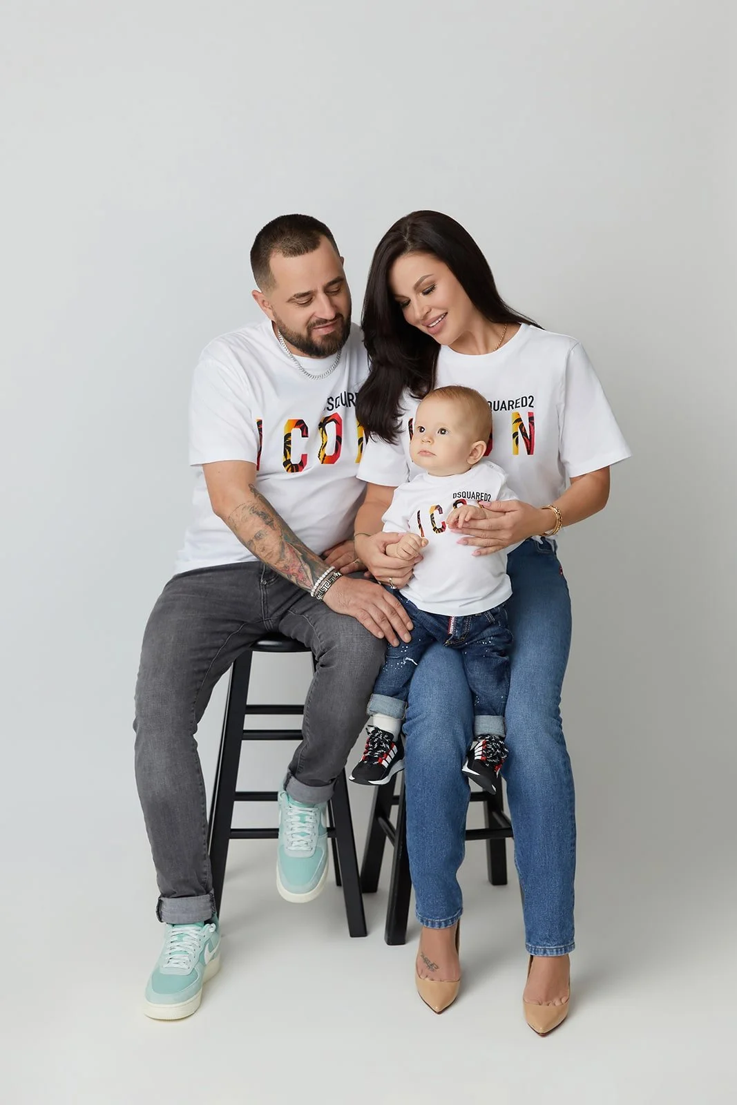 A family of three sitting on stools against a plain white background, all wearing matching white t-shirts with colorful printed text.