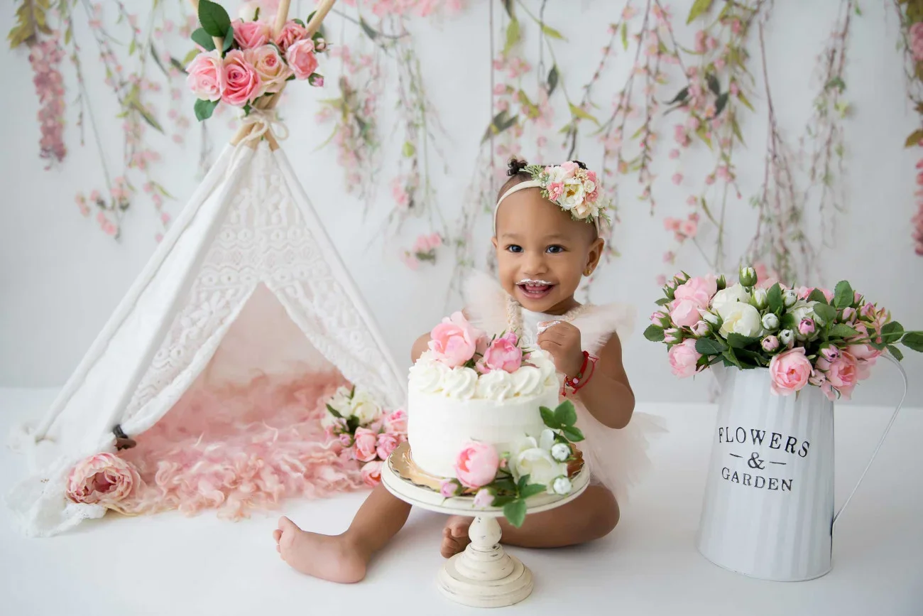 A young girl celebrating her birthday with a cake decorated with pink and white flowers, sitting next to a white flower vase and a pink floral teepee, in a pastel-colored room with pink floral decorations.