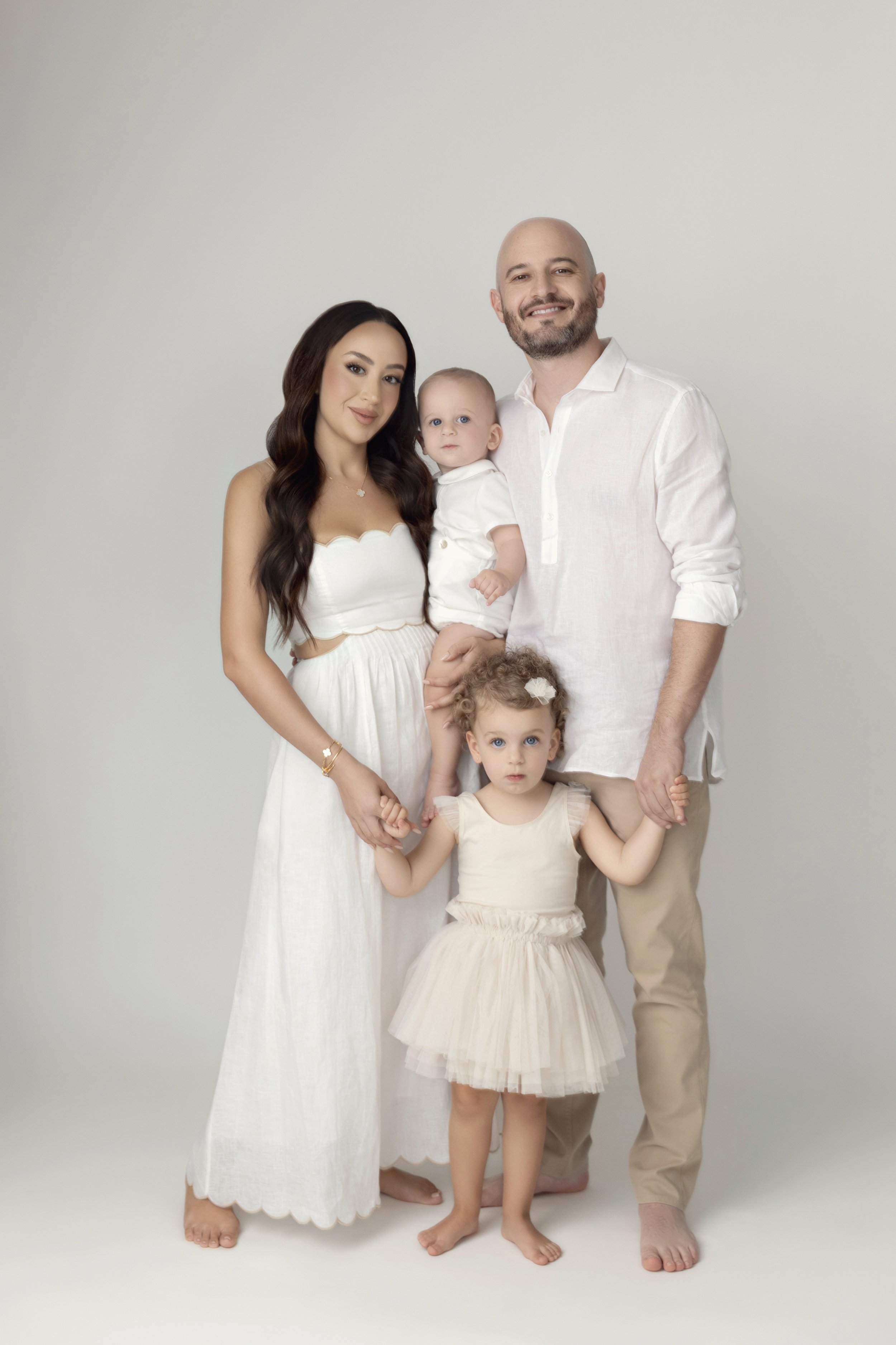 A family of four standing together against a plain white background, holding hands, dressed in light-colored clothing.