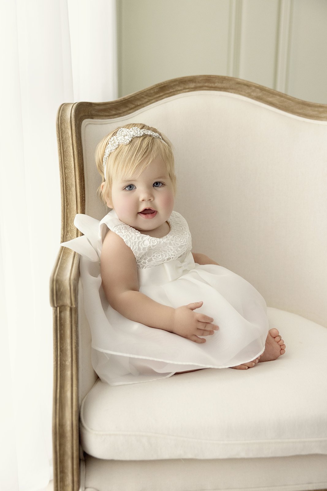 A young girl with blonde hair and blue eyes sitting on a cream-colored vintage-style couch, wearing a white dress with lace details and a decorative headband.
