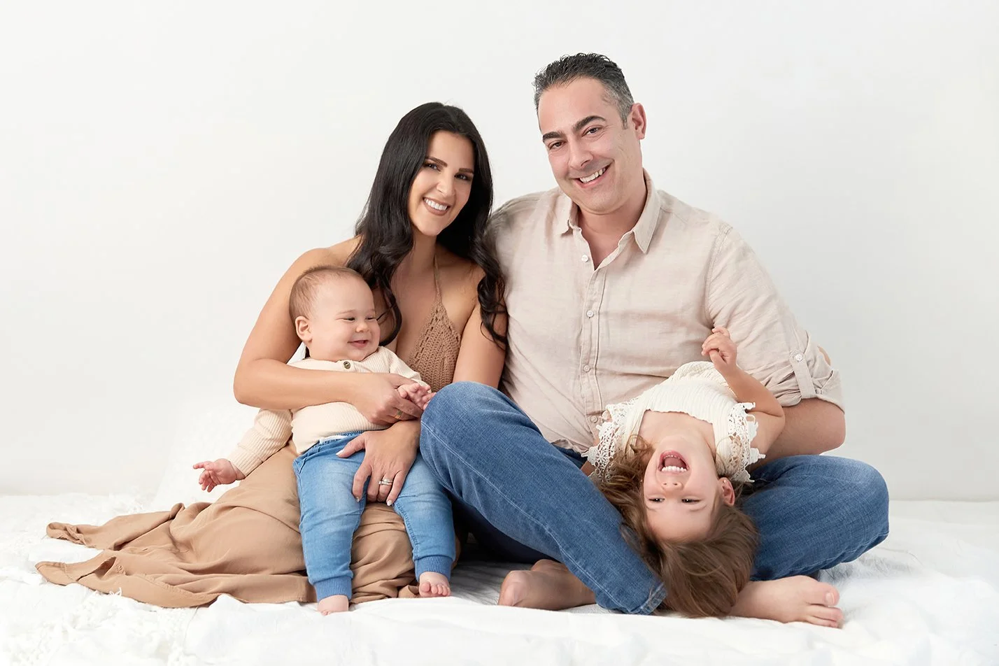 Family of four sitting on a white blanket, smiling and playing together, with a plain white background.