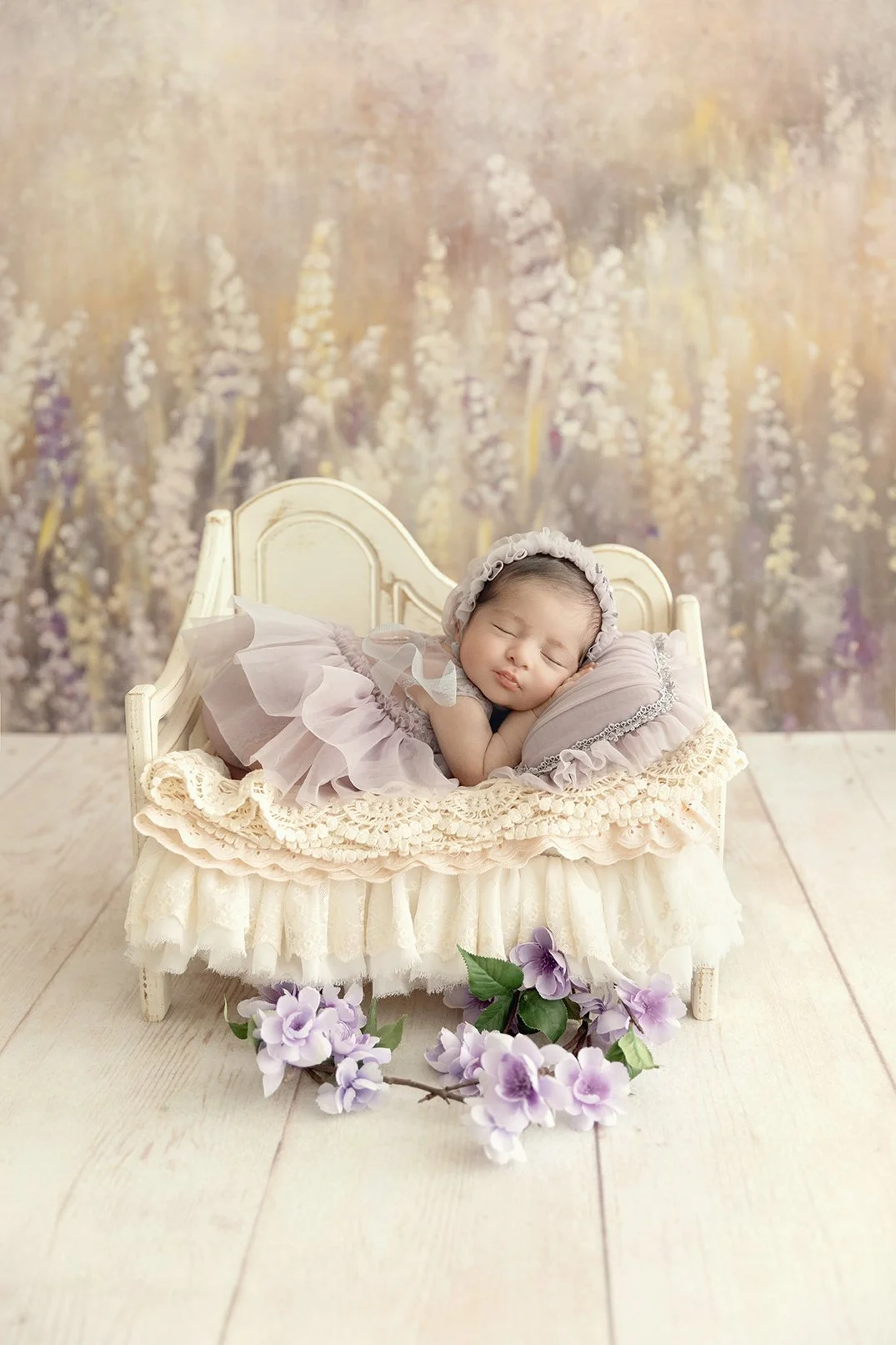 A sleeping baby girl lying on a miniature bed with floral decorations, dressed in a pink tutu and bonnet, with purple flowers in front of the bed and a blurred floral background.