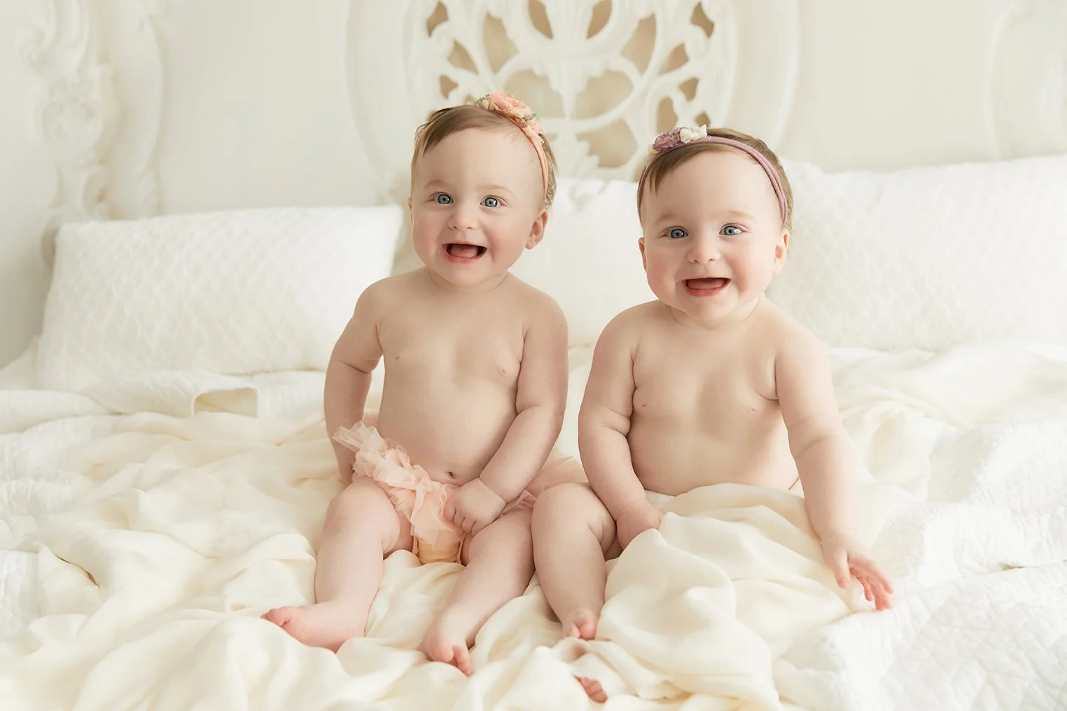 Two young babies sitting on a white bed, smiling and looking at the camera, each wearing a pink headband with a flower and light pink ruffled cloth around their waist.