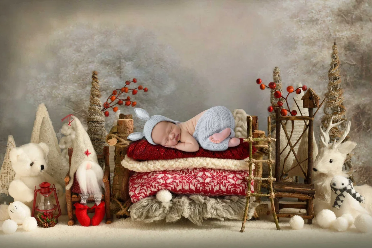 A sleeping baby lying on a decorated Christmas-themed setup with plush trees, a stuffed reindeer, a snowman, and a Santa figure surrounded by festive decorations.