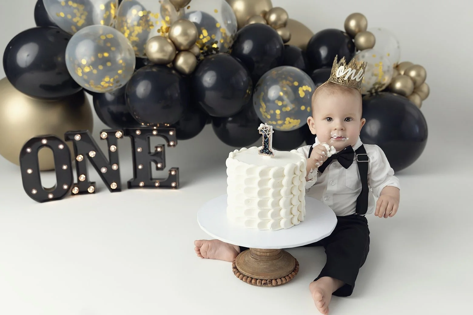 Baby boy celebrating first birthday, sitting near a white cake with a number one candle, wearing a birthday crown, surrounded by black, gold, and clear balloons, with a black illuminated "ONE" sign in the background.
