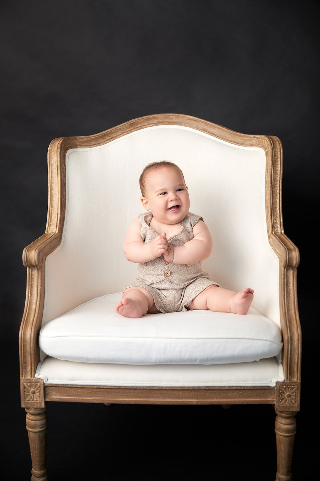 A smiling baby sitting on a vintage white armchair with wooden trim, against a dark background.
