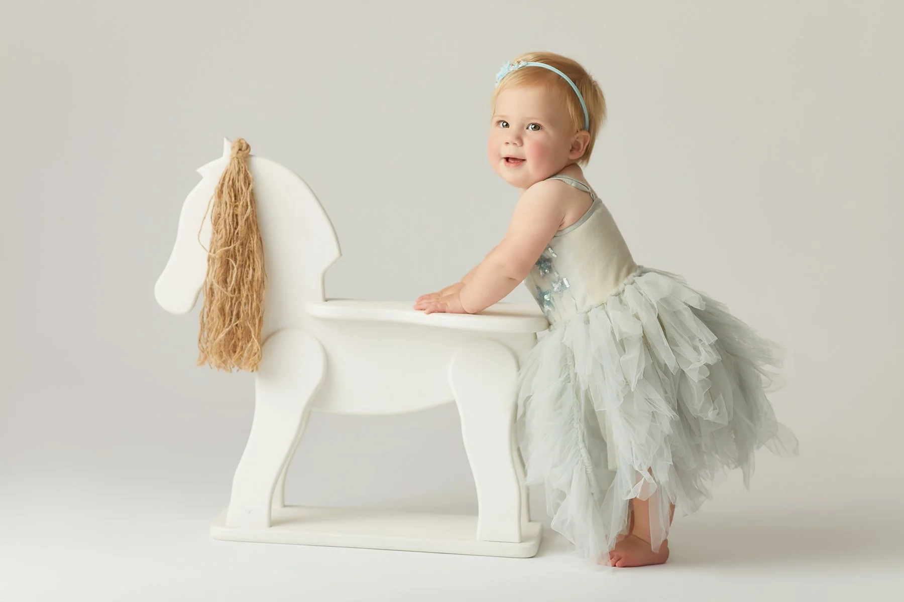 A smiling toddler girl with blonde hair and a blue headband, wearing a fancy, ruffled, silver dress, standing barefoot and leaning on a white wooden horse-shaped toy or decoration.