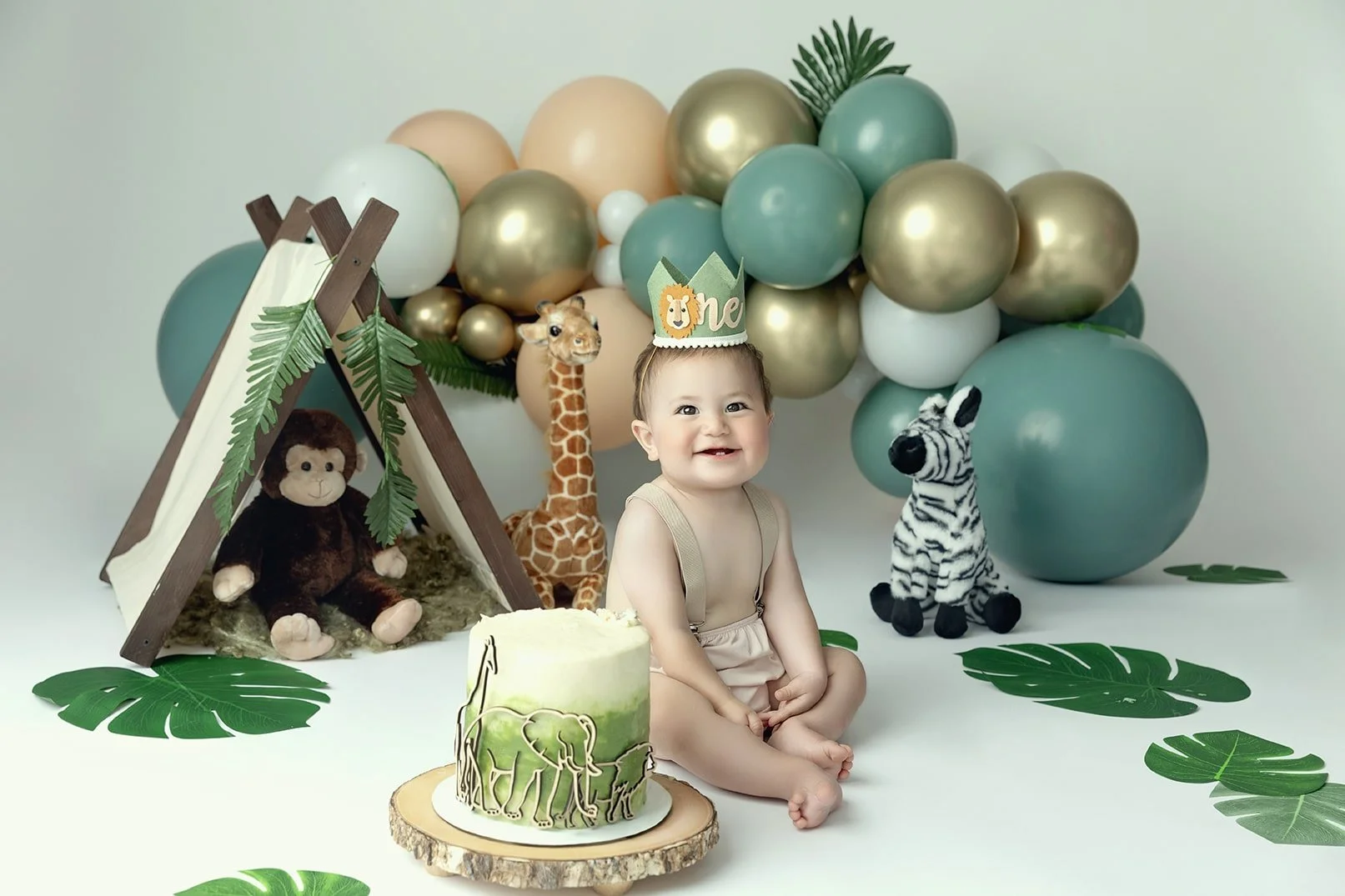 Baby boy sitting on floor in jungle-themed birthday setup with elephant cake, lion crown, and stuffed jungle animals, including giraffe, zebra, and monkey, with balloon backdrop and green leaves.