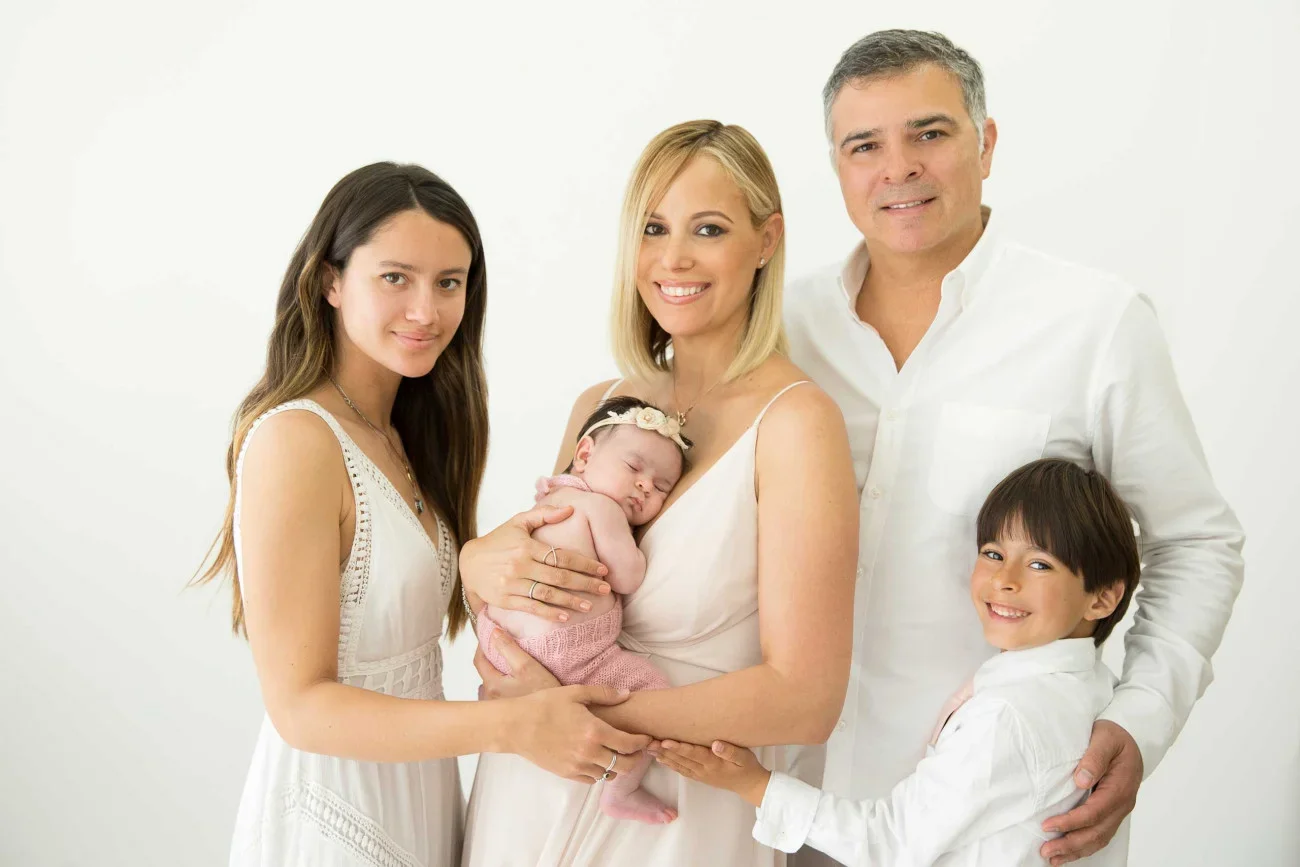 Family portrait of five members with a newborn baby, all dressed in white, standing against a plain white background, smiling at the camera.