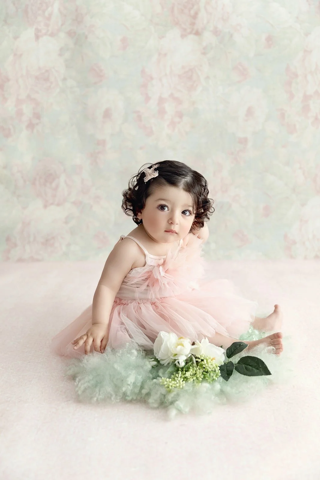 A young girl with curly dark hair wearing a pink dress and a hair clip, sitting on a soft surface with flowers and greenery in front of her, against a pastel floral background.