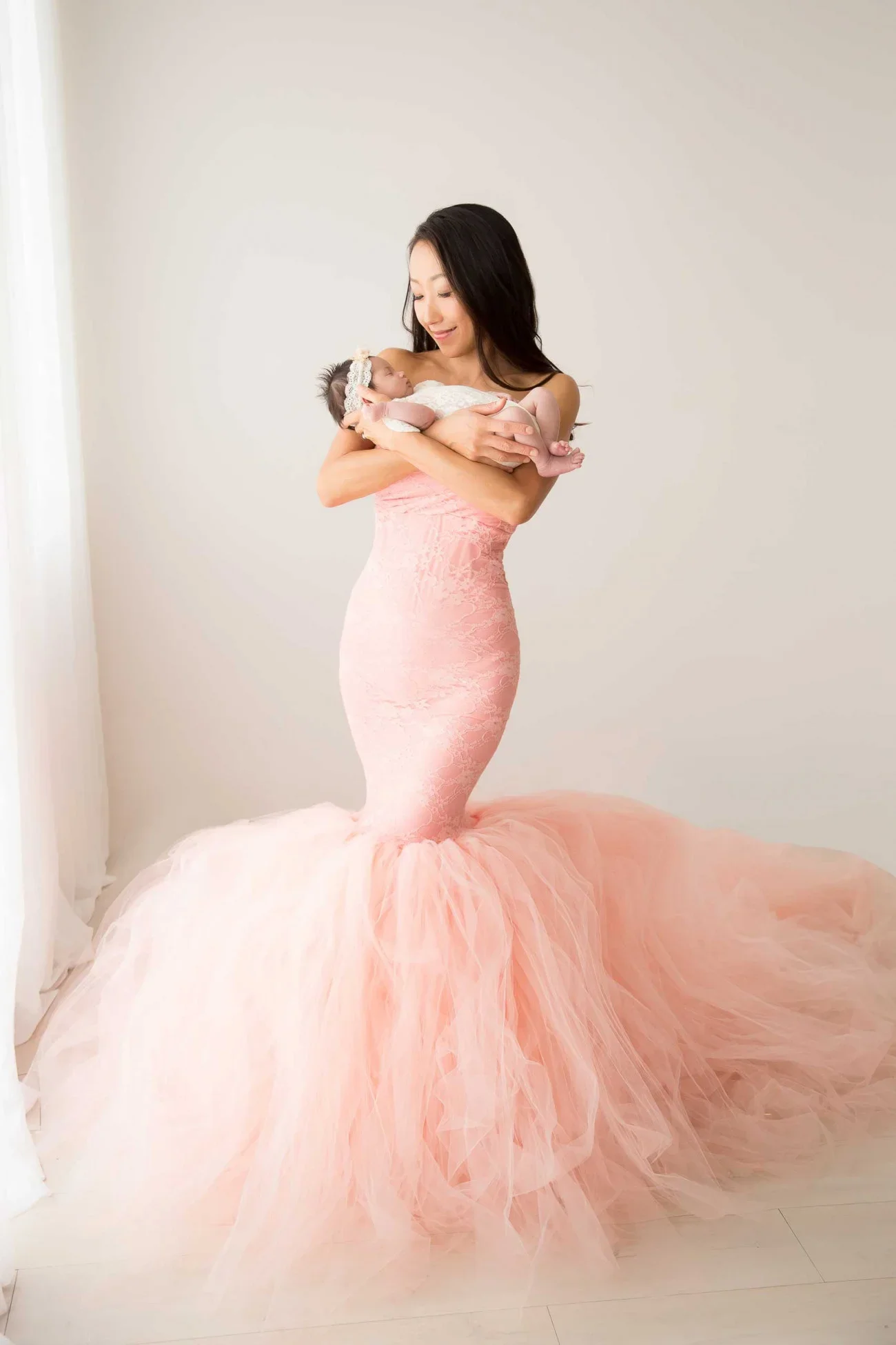 A woman in a pink, mermaid-style gown holding a small baby girl.