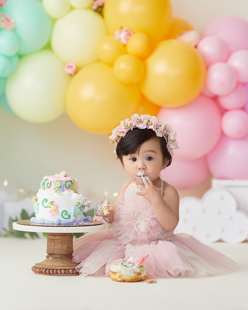 A baby girl in a pink dress and flower crown sits on the floor eating cake, with a birthday cake on a wooden stand next to her and colorful balloons in the background.