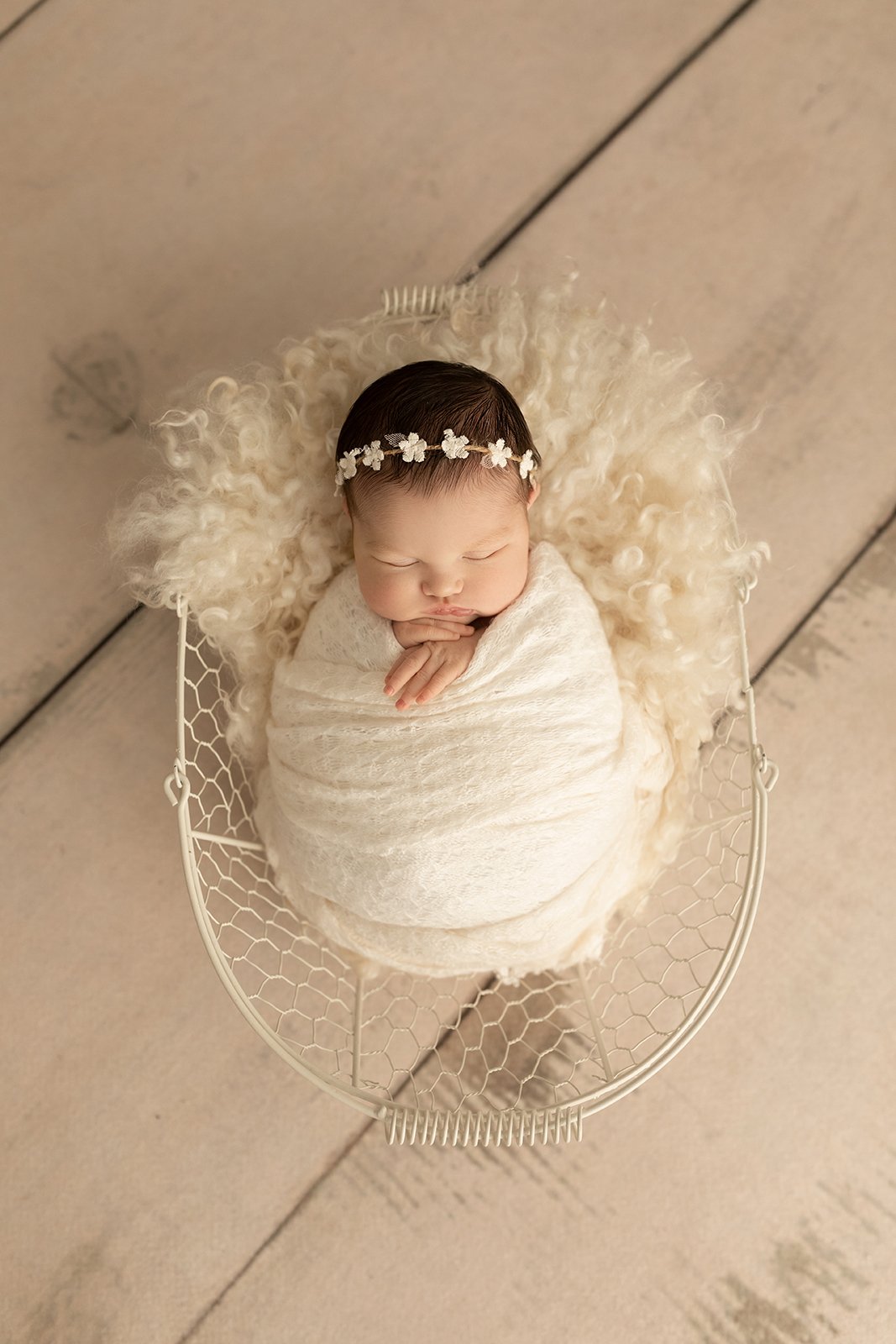 A sleeping newborn wrapped in a white swaddle, lying in a small wire basket with a cream-colored fluffy lining, on a wooden floor.