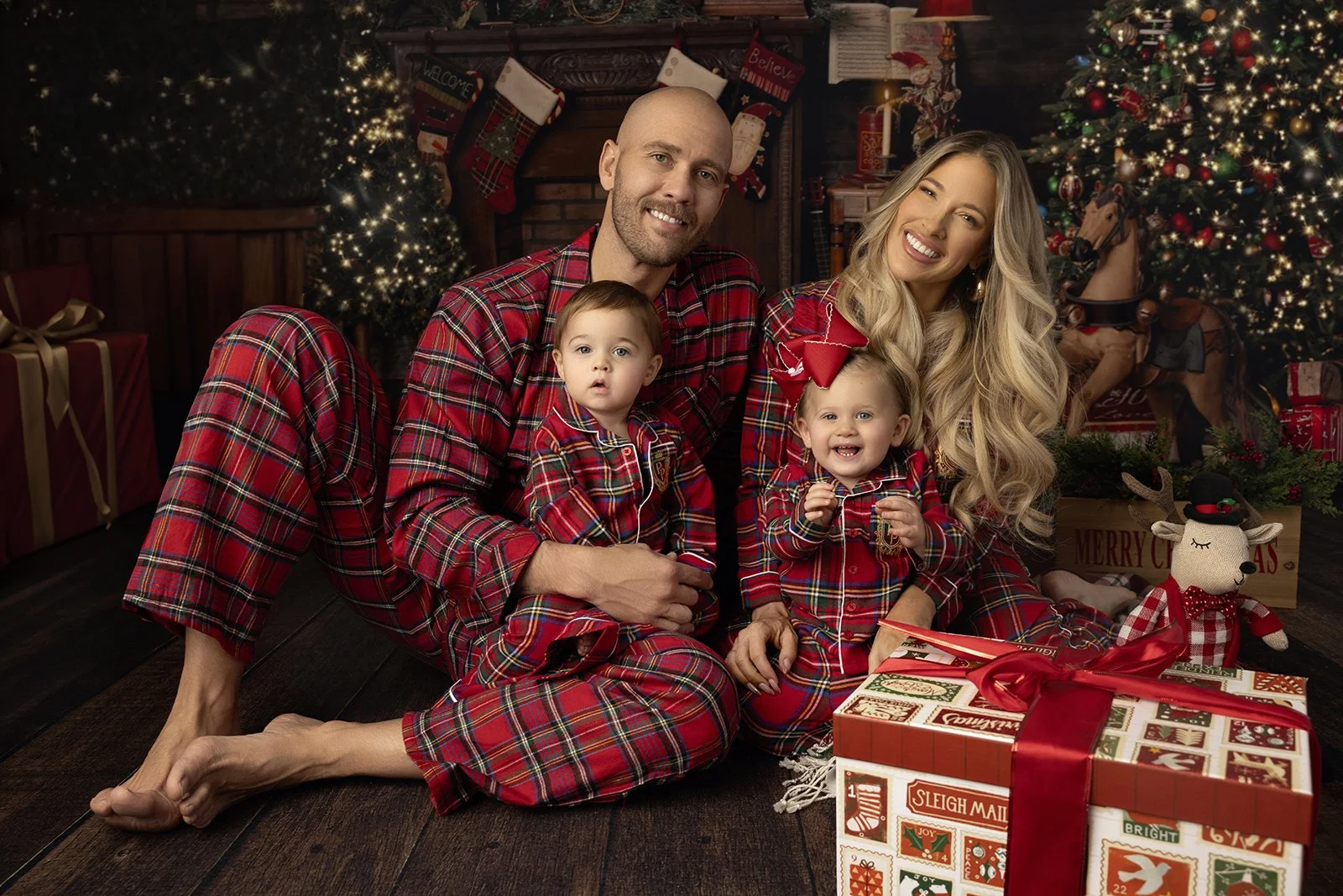 Family of four, dressed in matching red plaid pajamas, sitting on the wooden floor in front of a decorated Christmas fireplace with stockings, surrounded by wrapped gifts, a Christmas tree, and holiday decor.