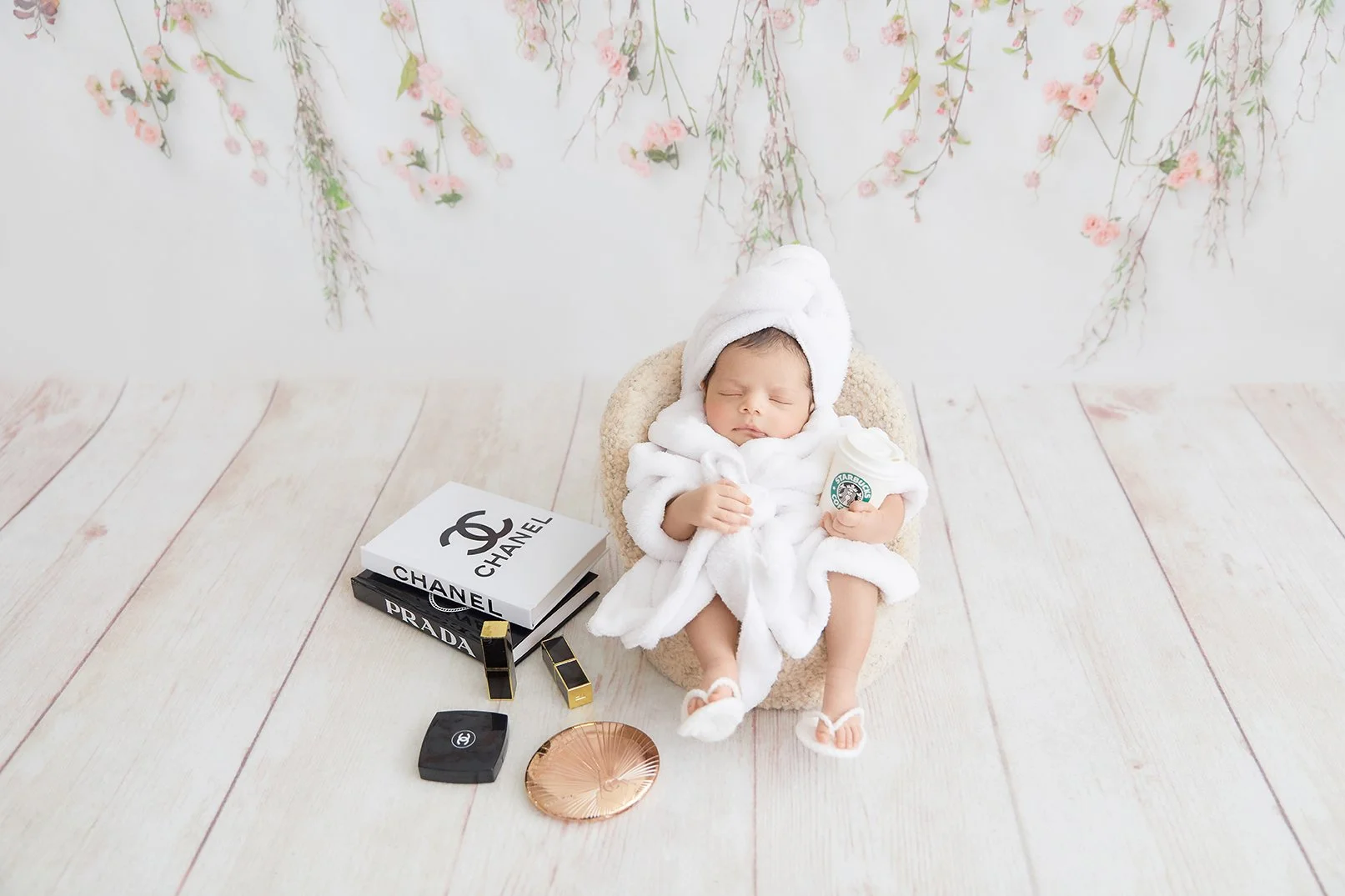Baby in a white robe and towel on head, holding a Starbucks cup, surrounded by luxury brand boxes and accessories, sitting on a plush chair on a wooden floor with floral backdrop.