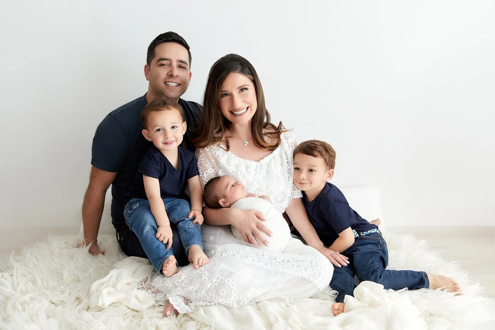 A family of five sitting together on a white furry rug against a white background. The mother is holding a newborn, with a father and two young children beside her, all smiling.